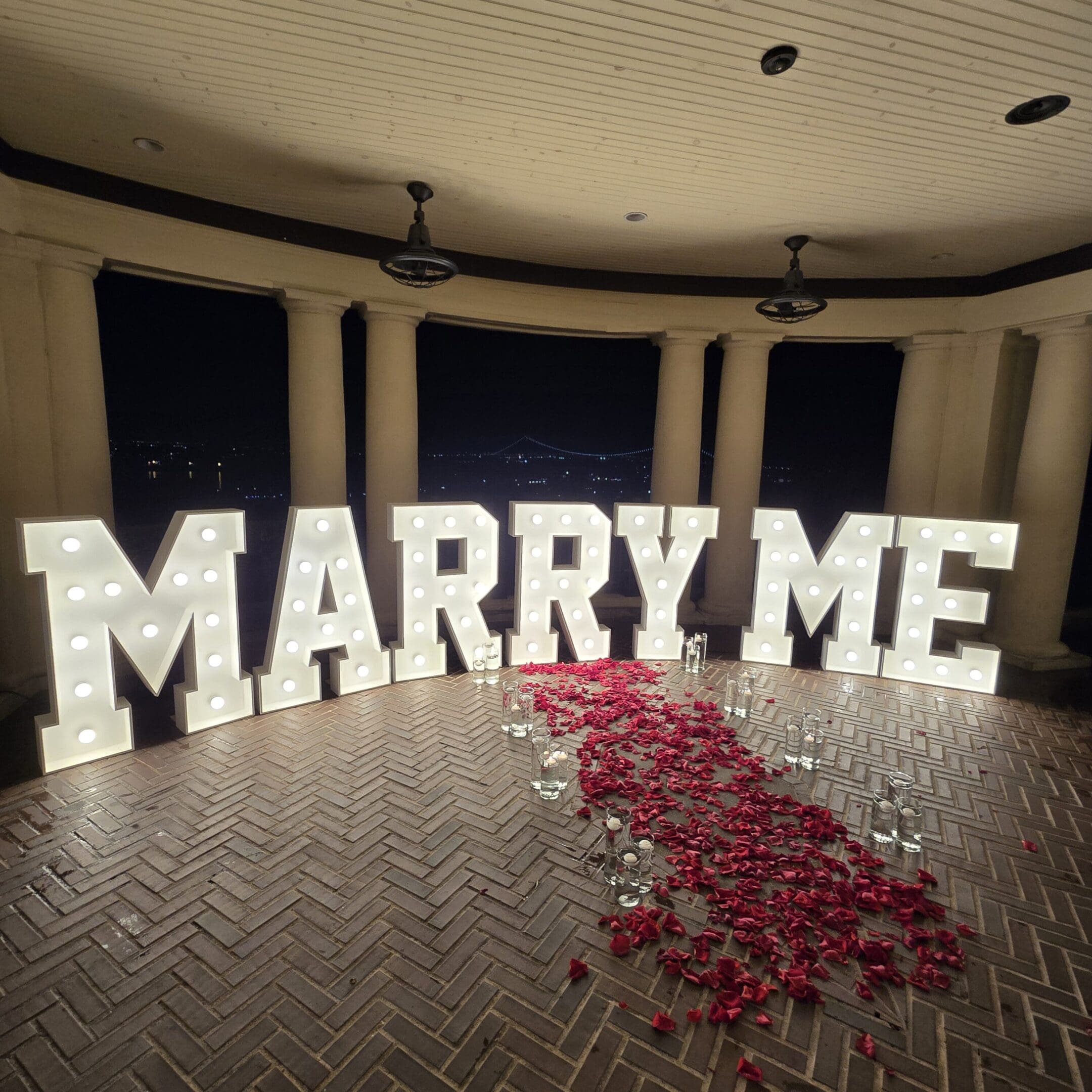 Illuminated 'MARRY ME' sign with rose petals on the floor for a proposal.