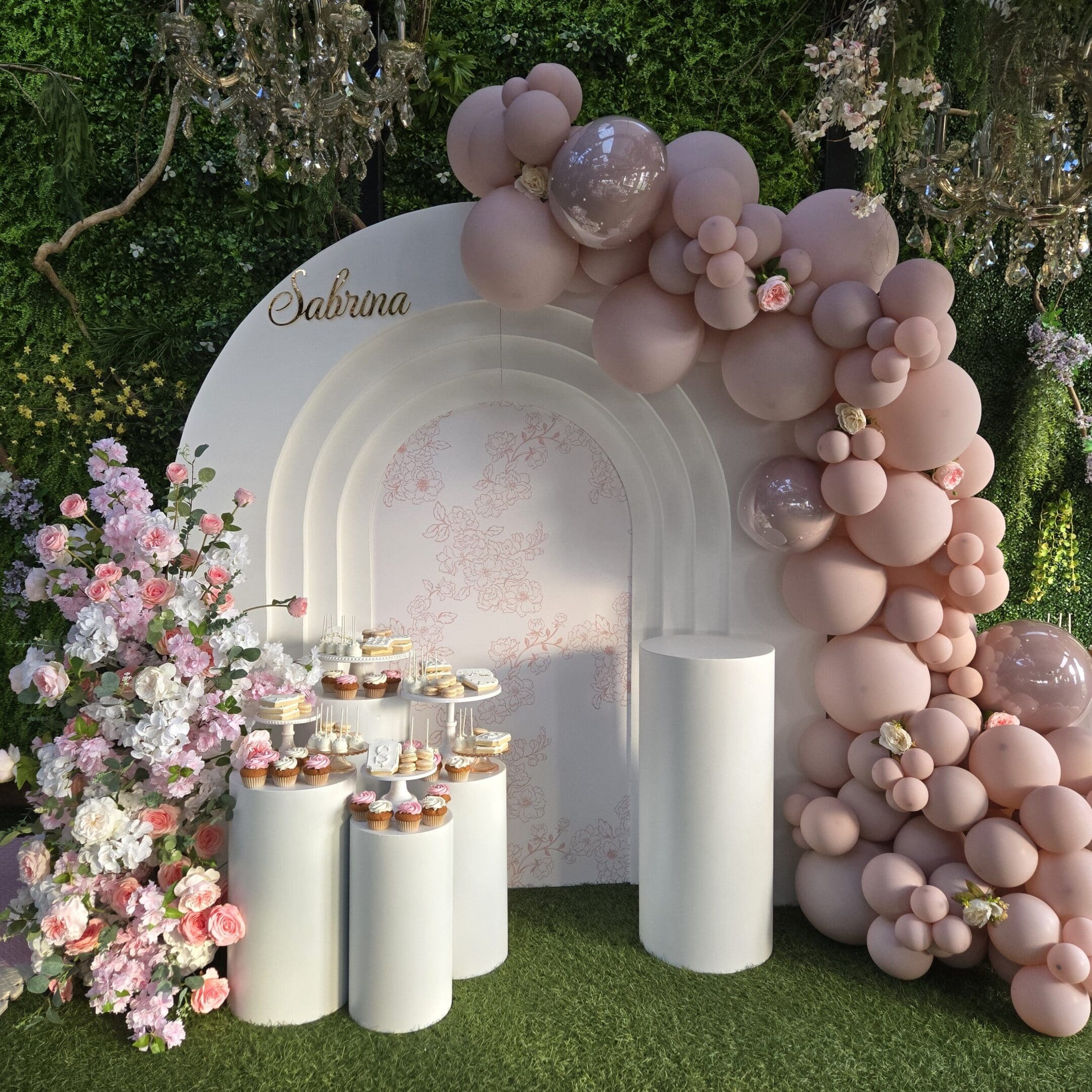 Elegant pastel balloon arch and floral decor with white pillars on mossy ground.